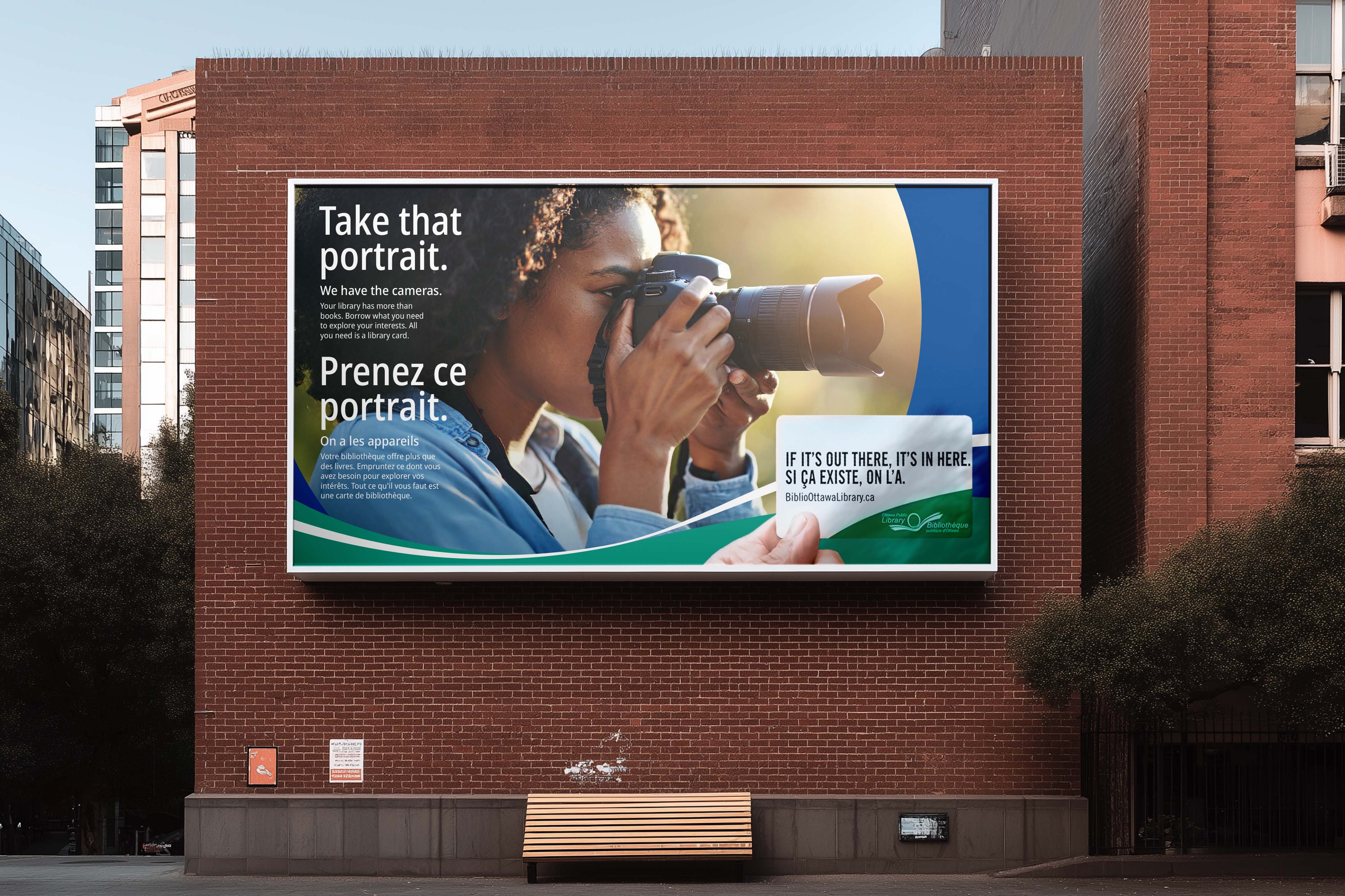 Billboard on a brick wall: a woman shooting with a big DSLR. Take that portrait. We have the cameras. Prenez ce portrait. On a les appareils. A hand holds out an Ottawa Public Library card.