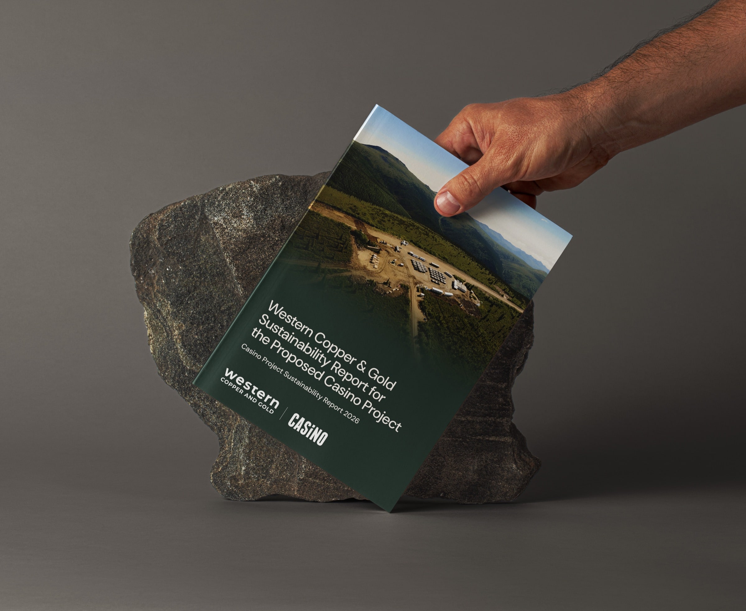 A hand holding the Western Copper and Gold sustainability report cover against a large rock.